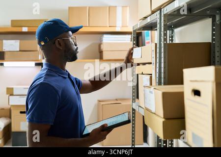 Black man young adult organizing cardboard boxes on storage shelf while holding digital tablet in hand, working in warehouse or stockroom, focusing on inventory management tasks Stock Photo