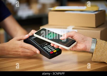 Caucasian young adult woman holding smartphone making contactless payment using point of sale terminal while another assisting with transaction in retail environment Stock Photo