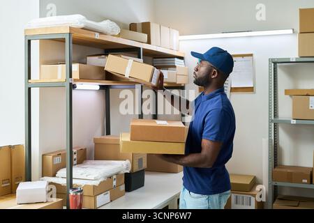 Young adult Black man organizing cardboard boxes on metal shelves in warehouse storage room, standing and reaching for package while wearing blue cap and polo shirt, surrounded by parcels Stock Photo