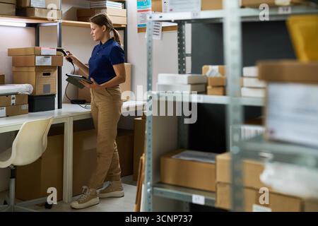 Young adult Caucasian woman standing in warehouse scanning packages with barcode scanner while holding digital tablet, surrounded by shelves filled with cardboard boxes and parcels Stock Photo