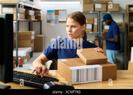 Caucasian young adult woman working at shipping desk holding cardboard boxes and using computer while Black man in background sorting packages in warehouse environment Stock Photo