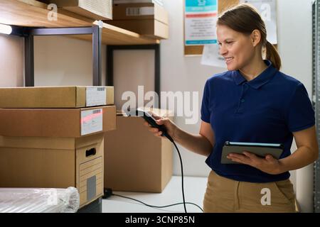 Caucasian young adult woman scanning cardboard boxes with barcode scanner while holding digital tablet in warehouse storage room, smiling and focusing on inventory management process Stock Photo
