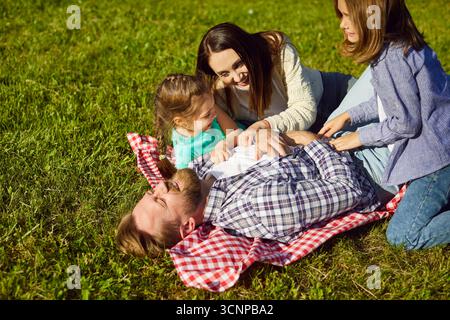 Happy family playing, father lying playfully on picnic blanket in sunny green park, tickled  Stock Photo