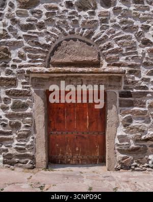 Weathered wooden door set in a rustic stone wall with an arched lintel. Irregular stones and textured mortar create a historical architectural background Stock Photo