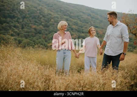 A family of three holds hands while walking through a golden meadow. The mother wears a pink blouse, and the daughter is in a light shirt. The scenic Stock Photo