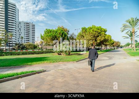 Man in suit carrying briefcase walks along a sunny city park pathway Stock Photo