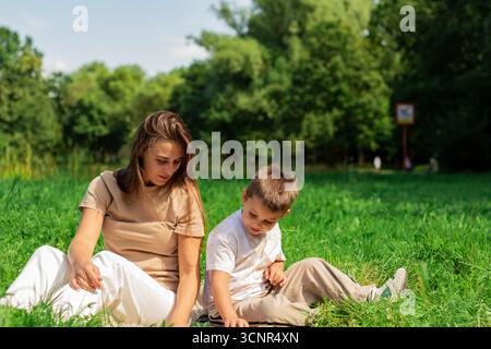 Mother helping her small son reading book about cars and transformers spending time on nature sitting on grass on summer day. Stock Photo