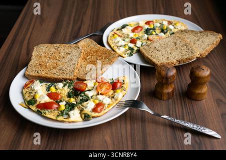 White plate with omelette, cherry tomatoes, spinach, cheese, and toast on wooden table, healthy homemade breakfast concept. Stock Photo