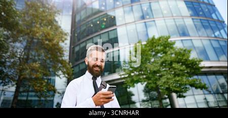 Handsome businessman in front of buildings in business district, looking at phone. Stock Photo