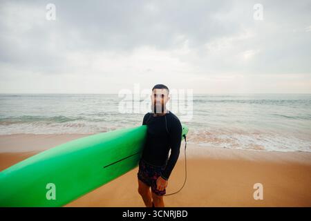 Young  bearded man with surfboard standing near a beach. Man with surfing board outdoors on a summer day. Stock Photo