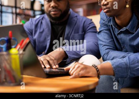Young remote workers in their living room collaborate on a project, using teamwork and digital online tools to enhance efficiency and productivity. Solving freelancing tasks at the coffee table. Stock Photo