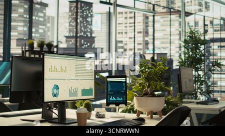 Modern workspace with analytical data paperwork documents in bright office environment with potted plants. Tablet on desk with digital statistics charts displayed on screen, panning shot Stock Photo