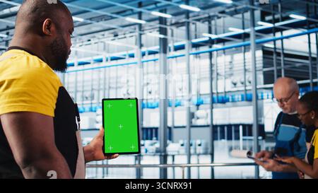 Smart factory expert using green screen tablet to test automated systems and ensure reliability. Worker uses chroma key device to identify bottlenecks and optimize gear based on sensors data, camera A Stock Photo