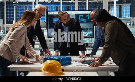 Factory management looking over schematics, working together to expand production. Industrial plant executives doing brainstorming, discussing over blueprint of facility layout, camera A Stock Photo