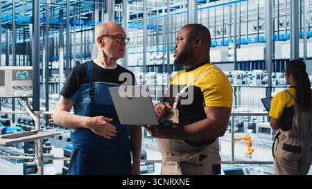 Smart factory senior engineer teaching new trainee to manage robotic arms. Industry 4.0 industrial plant team leader and intern controlling automated machinery, ensuring workflow continuity, camera A Stock Photo