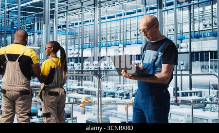 Industry 4.0 industrial plant senior engineer automating robotic machines using laptop software. Experienced technician implementing AI features in smart factory using notebook, camera A Stock Photo