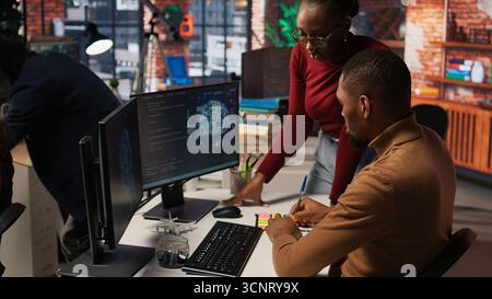 Teamworking startup engineers writing message on sticky notes while coding advanced AI algorithm. Coworkers in artificial intelligence Silicon Valley workspace leaving memo on PC screen, camera A Stock Photo