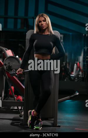 blonde woman wearing a fitted black top and leggings stands confidently at the gym. She poses near weightlifting equipment showcasing her dedication to fitness and health. Stock Photo