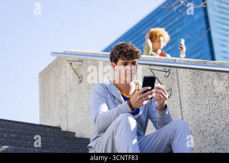 Man sitting on steps with smartphone and earbuds in plaza, woman by railing checking phone Stock Photo