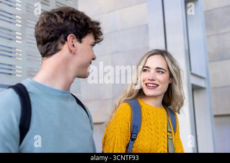 Diverse classmates walking and chatting outside stone facade with backpacks near metal panel Stock Photo