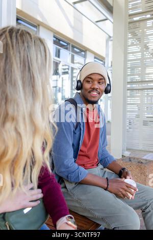 Diverse student friends sitting on wooden bench at bus stop holding coffee cup, green backpack Stock Photo