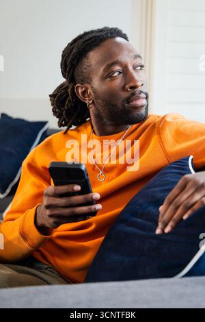 African American man wearing orange sweatshirt, necklace sitting on sofa at home holding smartphone Stock Photo