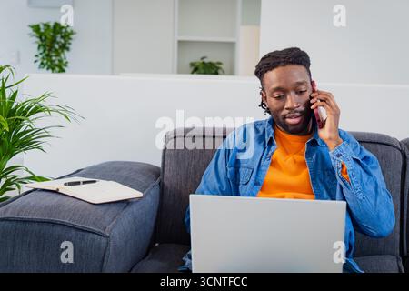 African American man talking on phone, typing on laptop on sofa in home office, copy space Stock Photo