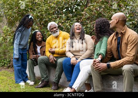 Diverse family gathering on wooden log bench in park wearing autumn sweaters sporting smartwatch Stock Photo
