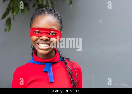 African American girl wearing red mask and blue scarf, standing outdoors against gray wall, smiling Stock Photo