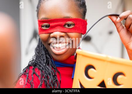 African American girl smiling and holding jack-o'-lantern candy bucket on porch wearing red mask Stock Photo