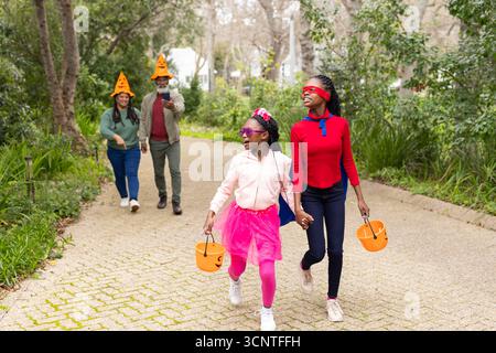Diverse family walking pathway amid autumn leaves holding pumpkin buckets and filming on smartphone Stock Photo