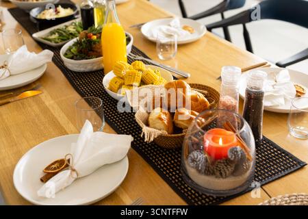 Black runner is sitting on wooden table with bread rolls, corn, orange juice bottle, lit candle Stock Photo