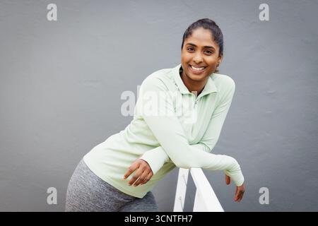 South Asian woman in athletic wear leaning against white metal railing on terrace smiling at camera Stock Photo