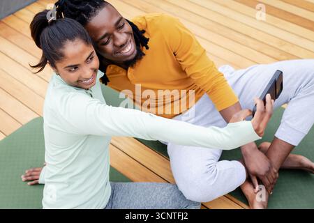 Diverse couple taking selfie on green yoga mats on wooden deck holding smartphone Stock Photo
