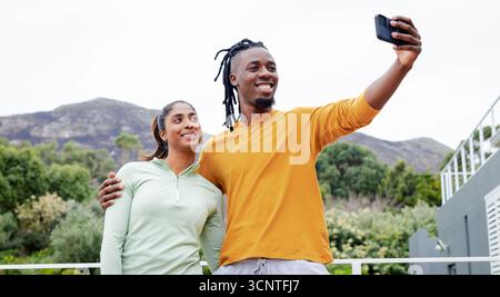 Diverse couple in activewear taking selfie on balcony overlooking hills with smartphone and railing Stock Photo