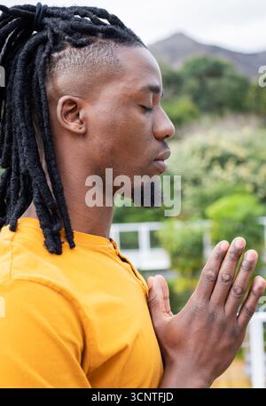African American man praying on balcony wearing mustard yellow shirt under overcast sky, copy space Stock Photo