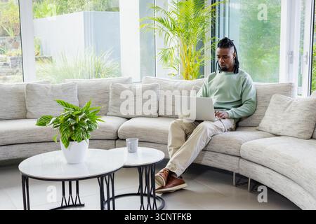 African American man sitting on sectional sofa typing on silver laptop in living room, copy space Stock Photo