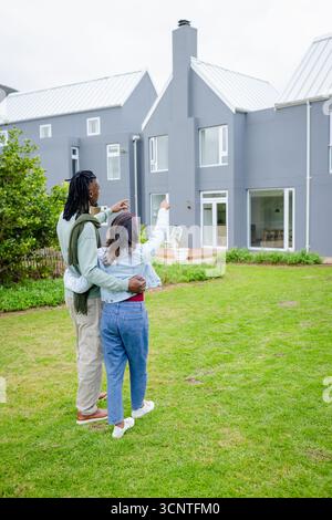 Diverse couple standing on green lawn pointing toward modern grey house with white metal roof Stock Photo