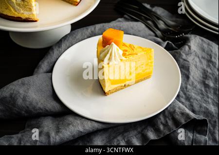 Slice of Mango Mousse Cake on a Plate Angled View: Piece of swirled mousse cake with with a stack of plates, forks, and cake stand in the background Stock Photo