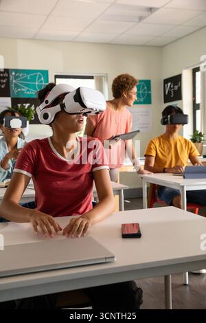 Diverse classmates wearing vr headsets using laptops while teacher with tablet in classroom Stock Photo