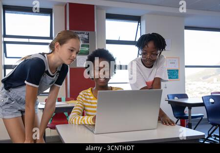 Diverse child classmates gathering around laptop on desk in classroom Stock Photo