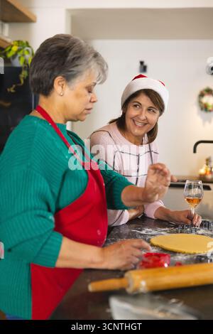 Mother and daughter wearing apron pressing cookie cutter into dough while holding wine in kitchen Stock Photo