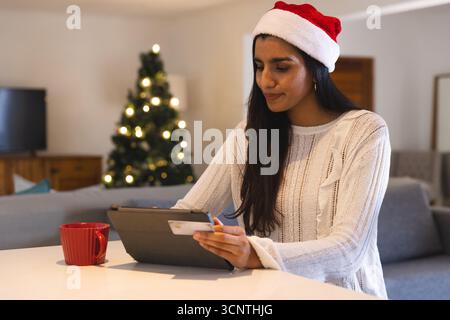 Indian woman sitting at home table using tablet and credit card near Christmas tree, copy space Stock Photo