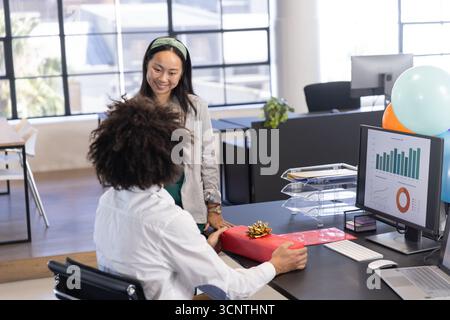 Diverse coworkers exchanging wrapped red gift box at desk with charts on screen in office Stock Photo