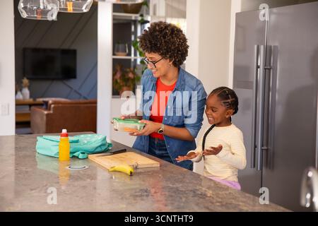 Hispanic mother with african american daughter packing lunch box on kitchen island with backpack Stock Photo