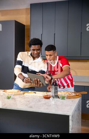 Diverse male friends preparing pizzas at marble kitchen island using tablet and wooden pizza boards Stock Photo