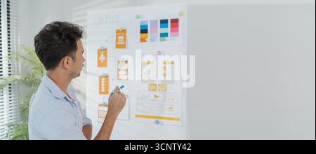 A young designer is working on a UX UI project, analyzing mobile app wireframes and choosing color palettes on a whiteboard. He focuses on brainstormi Stock Photo