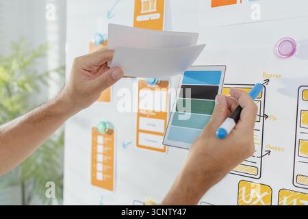 A young designer is working on a UX UI project, analyzing mobile app wireframes and choosing color palettes on a whiteboard. He focuses on brainstormi Stock Photo