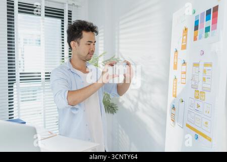 A creative designer works on UX/UI planning, analyzing sketches and prototypes on a whiteboard while using a laptop. The process shows design thinking Stock Photo