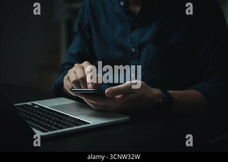 A man is using his cell phone while sitting at a desk with a laptop. The man is focused on his phone, possibly texting or browsing the internet. Conce Stock Photo